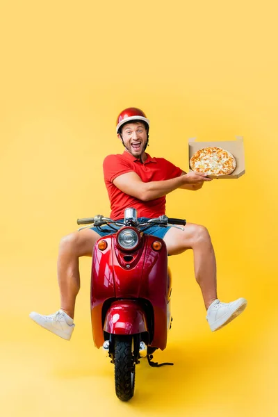 Excited delivery man in helmet riding scooter and holding pizza in carton box on yellow — Stock Photo