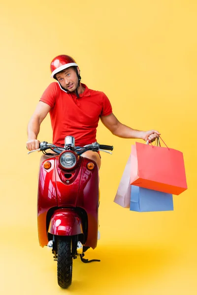 Livreur dans casque équitation scooter et smartphone parler tout en tenant des sacs à provisions sur jaune — Photo de stock