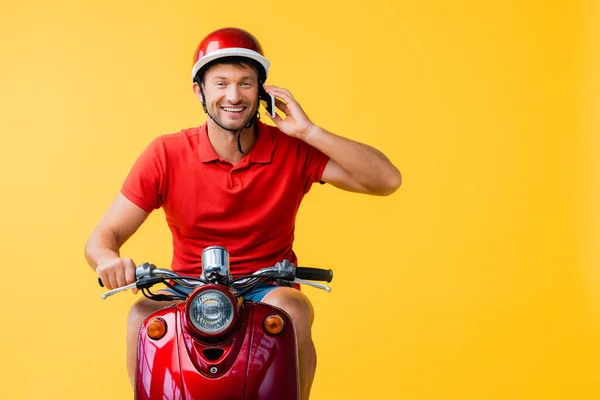 Задоволений чоловік доставки в шолом їде скутер і розмовляє на смартфоні на жовтому — стокове фото