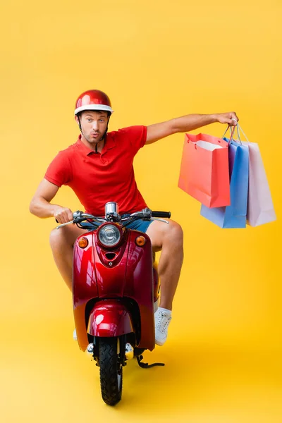 Удивленный курьер в шлеме катается на скутере и держит сумки для покупок на желтом — стоковое фото