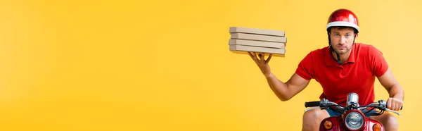 Serious delivery man in helmet riding scooter and holding carton pizza boxes on yellow, banner — Stock Photo