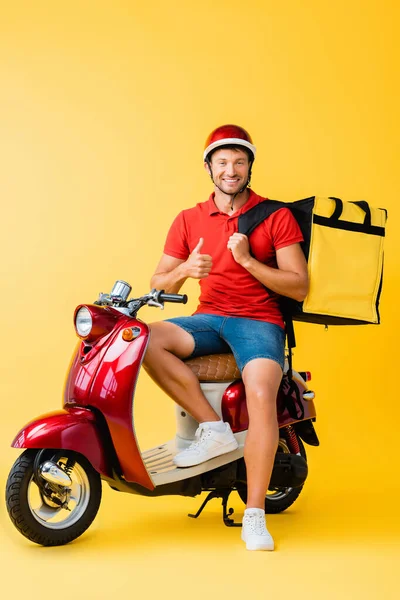 Happy delivery man with backpack sitting on red scooter and showing thumb up on yellow — Stock Photo