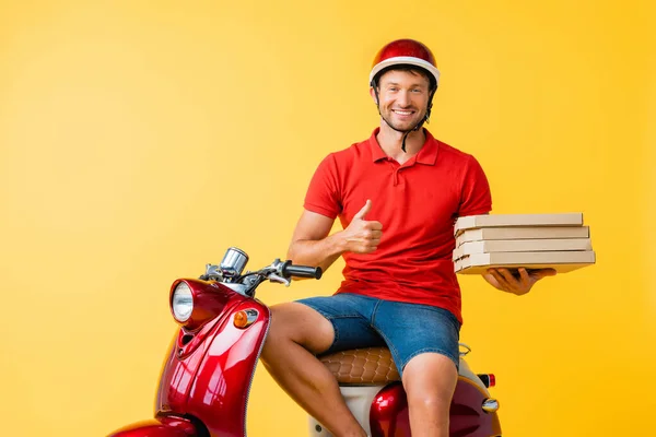 Felice uomo di consegna in casco tenendo scatole di pizza mentre mostra pollice verso l'alto vicino scooter sul giallo — Foto stock