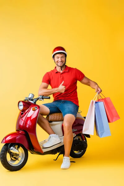 Happy delivery man in helmet sitting on scooter and holding shopping bags while showing thumb up on yellow — Stock Photo