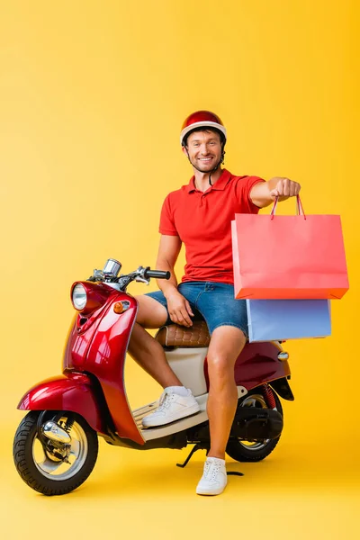 Happy delivery man in helmet sitting on scooter and holding shopping bags on yellow — Stock Photo