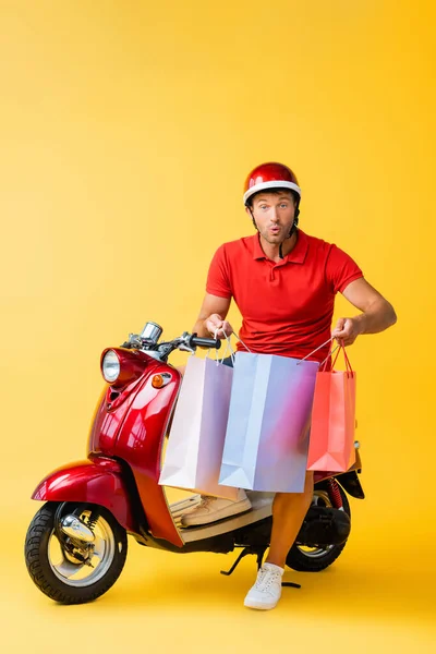 Homme de livraison choqué dans le casque assis sur scooter et tenant des sacs à provisions sur jaune — Photo de stock