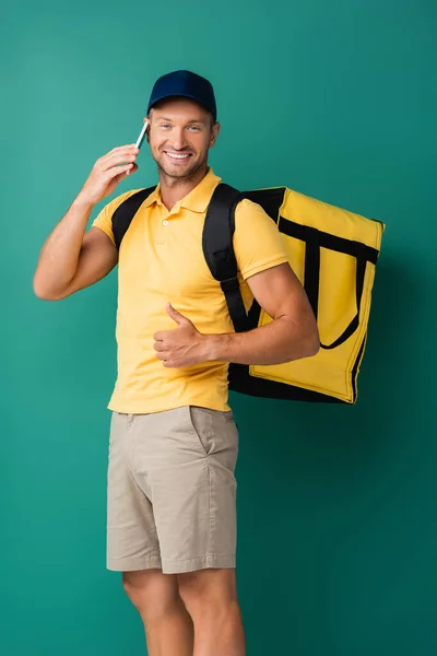 Alegre repartidor hombre llevando mochila amarilla y hablando en el teléfono inteligente en azul - foto de stock