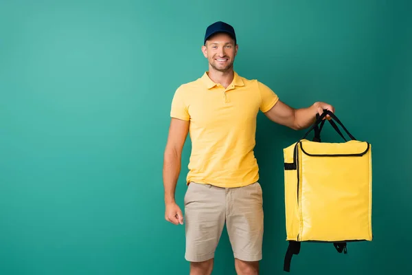 Cheerful delivery man carrying yellow backpack and smiling on blue — Stock Photo