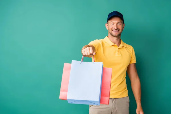 Cheerful delivery man with outstretched hand carrying shopping bags on blue — Stock Photo