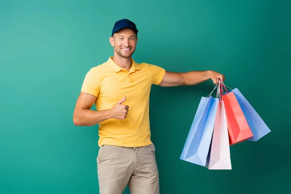 Homem de entrega alegre em cap carregando sacos de compras e mostrando polegar para cima no azul — Fotografia de Stock