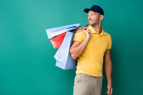 Alegre repartidor hombre en gorra llevando bolsas de compras y mirando hacia otro lado en azul - foto de stock