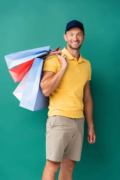 Fröhlicher Auslieferer in Mütze trägt Einkaufstüten auf blauem Grund — Stockfoto