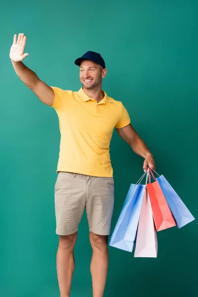 Cheerful delivery man in cap carrying shopping bags and waving hand on blue — Stock Photo