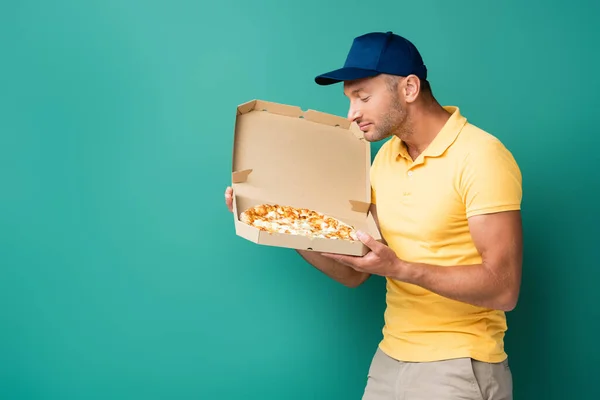 Complacido repartidor hombre oliendo sabrosa pizza en caja en azul - foto de stock