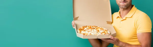 Обрезанный вид курьера, кусающего губы, держа вкусную пиццу в коробке на голубом, баннер — стоковое фото