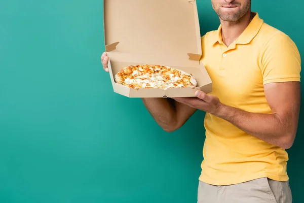 Vista recortada del repartidor hombre mordiendo labios mientras sostiene sabrosa pizza en caja en azul - foto de stock