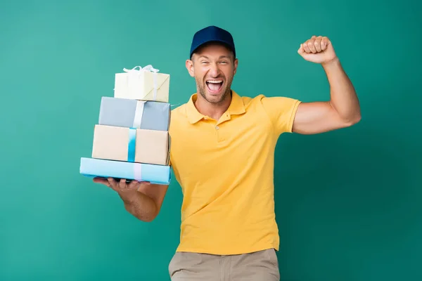 Uomo di consegna eccitato in cap tenendo avvolto regali su blu — Foto stock