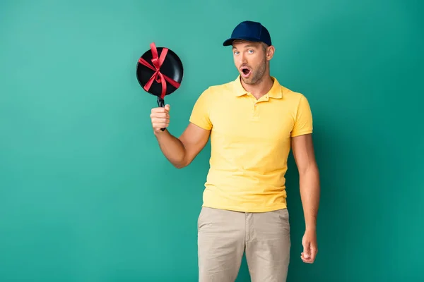 Shocked delivery man in cap holding frying pan with ribbon on blue — Stock Photo