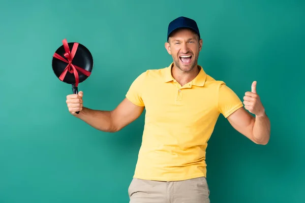 Excited delivery man in cap holding frying pan with ribbon on blue — Stock Photo
