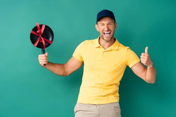 Aufgeregter Auslieferer in Mütze mit Bratpfanne und Schleife auf blauem Grund — Stockfoto