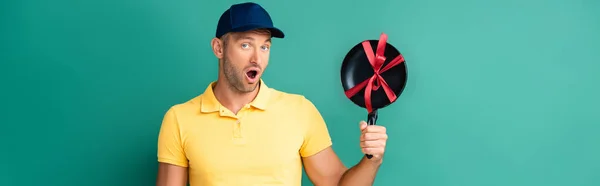 Shocked delivery man in cap holding frying pan with ribbon on blue, banner — Stock Photo