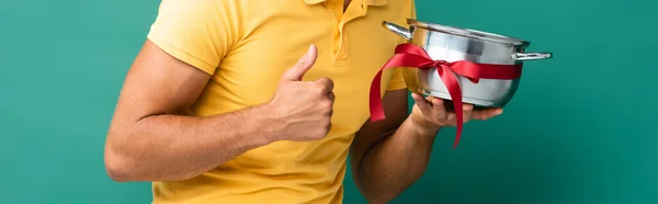 Cropped view of delivery man holding saucepan with ribbon and showing thumb up on blue — Stock Photo