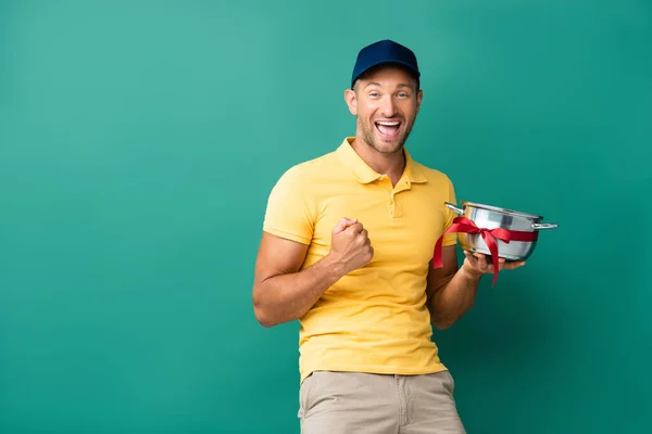 Smiling delivery man in cap holding saucepan with ribbon on blue — Stock Photo