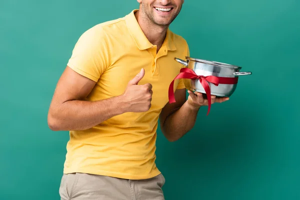 Cropped view of cheerful delivery man holding saucepan with ribbon and showing thumb up on blue — Stock Photo