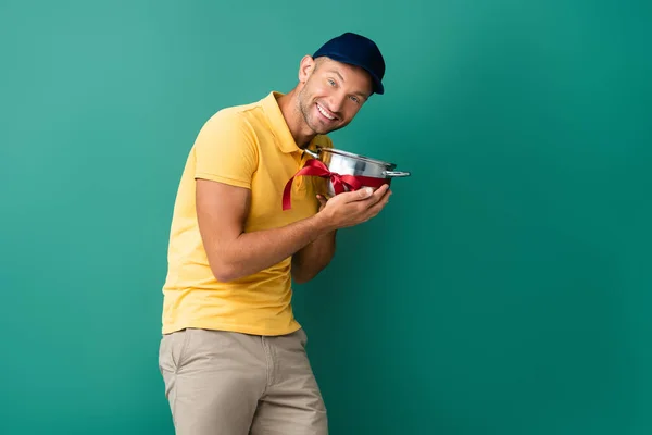 Cheerful delivery man in cap holding saucepan with ribbon on blue — Stock Photo