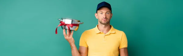 Delivery man in cap holding saucepan with ribbon on blue, banner — Stock Photo