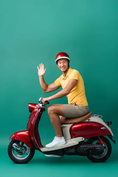 Felice uomo di consegna in casco cavalcando moto e agitando mano sul blu — Foto stock