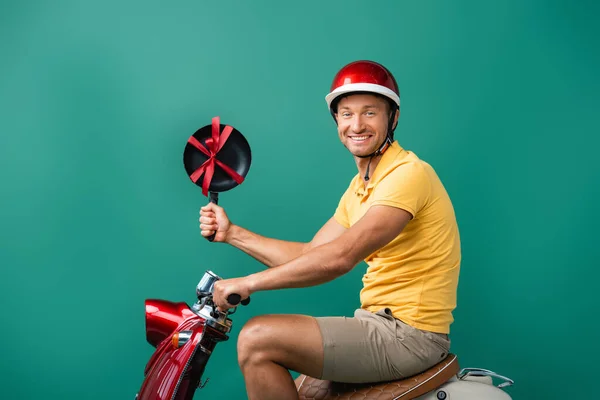 Fröhlicher Auslieferer im Helm, Motorrad fahrend und Bratpfanne mit Schleife auf blau haltend — Stockfoto