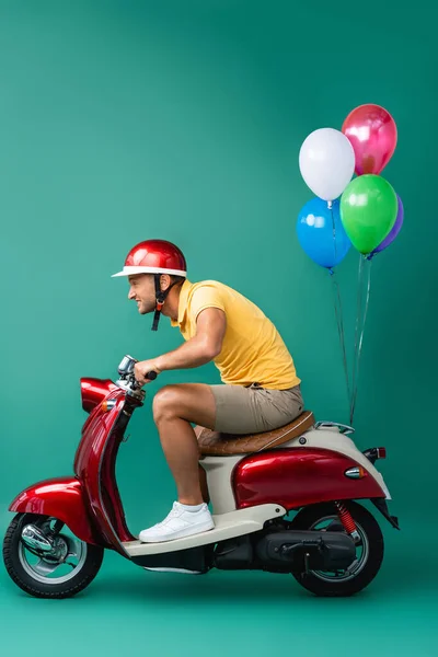 Vista laterale dell'uomo di consegna concentrato in casco cavalcando scooter rosso con palloncini su blu — Foto stock