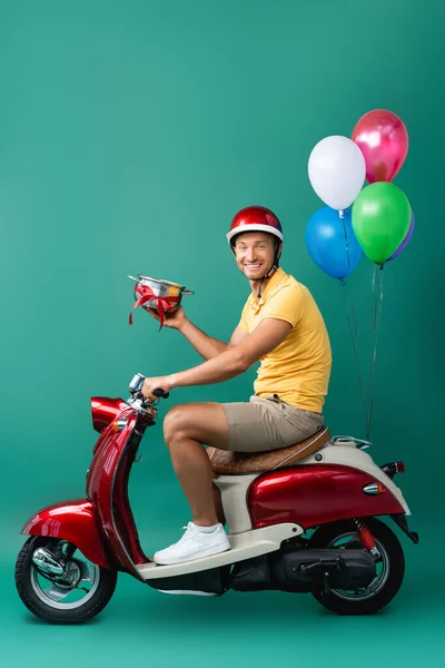 Feliz repartidor en casco sosteniendo cacerola con cinta mientras monta scooter con globos en azul - foto de stock