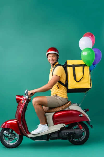 Heureux livreur avec sac à dos scooter équitation avec ballons sur bleu — Photo de stock