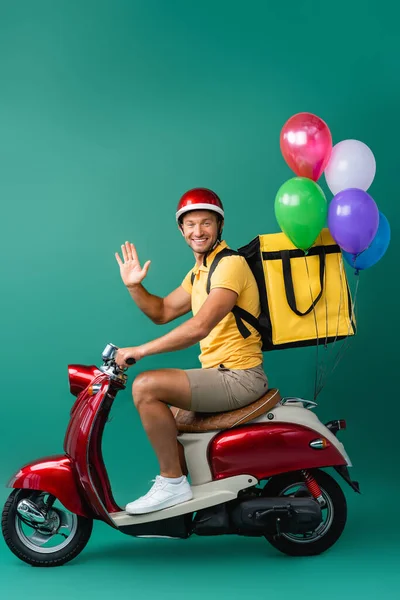 Happy delivery man with backpack riding scooter with balloons and waving hand on blue — Stock Photo