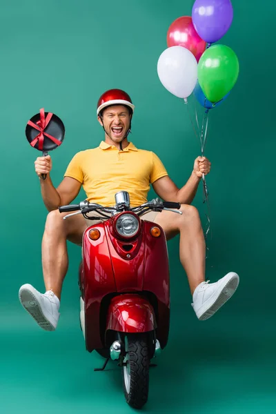 Excited delivery man in helmet riding scooter while holding balloons and new frying pan on blue — Stock Photo