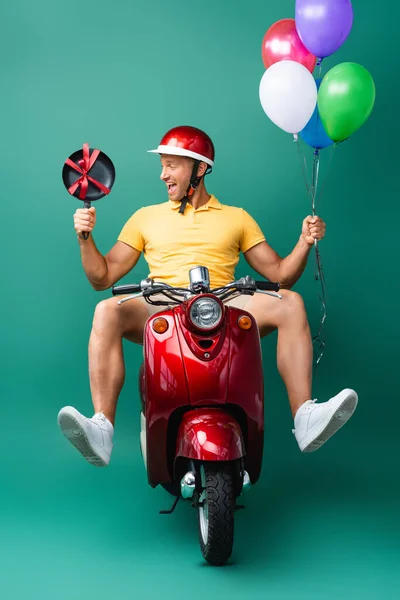 Heureux livreur en scooter équitation casque tout en tenant des ballons et une nouvelle poêle sur bleu — Photo de stock