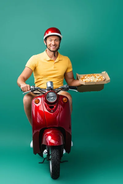 Heureux livreur en scooter d'équitation casque tout en tenant savoureuse pizza en boîte sur bleu — Photo de stock