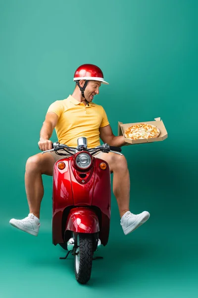 Excited delivery man in helmet riding scooter while holding tasty pizza in box on blue — Stock Photo