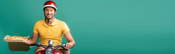 Hombre feliz de la entrega en el casco de montar scooter mientras que sostiene la pizza sabrosa en caja en azul, bandera - foto de stock