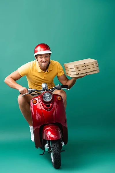 Чоловік доставки в шолом кусає губи під час їзди на скутері, тримаючи коробки для піци на синьому — стокове фото