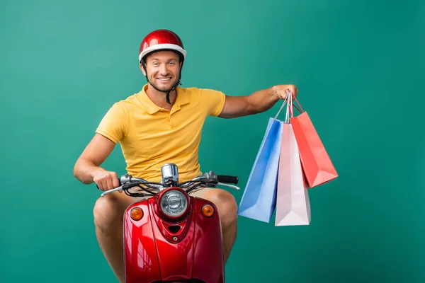 Heureux livreur en scooter d'équitation casque tout en tenant des sacs à provisions sur bleu — Photo de stock