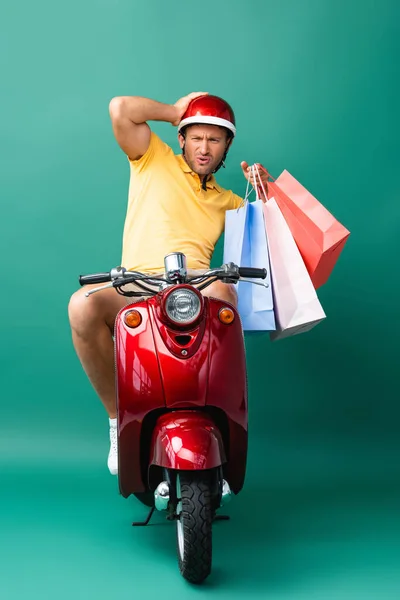 Homem de entrega chocado no capacete equitação scooter enquanto segurando sacos de compras em azul — Fotografia de Stock