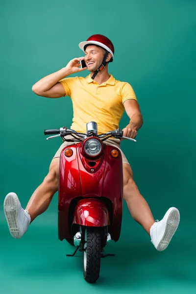 Cheerful delivery man in helmet riding scooter while talking on smartphone on blue — Stock Photo