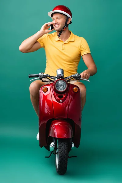 Щасливий чоловік доставки в шолом їде скутер під час розмови на смартфоні на синьому — стокове фото