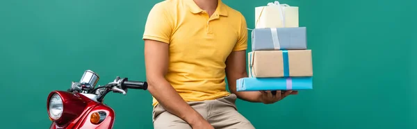 Partial view delivery man in helmet holding wrapped presents near scooter on blue, banner — Stock Photo