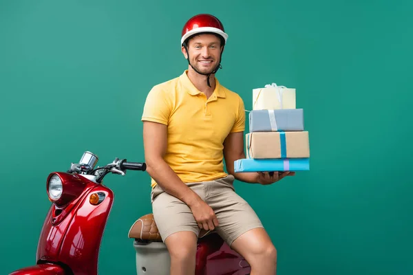 Happy delivery man in helmet holding wrapped presents near scooter on blue — Stock Photo