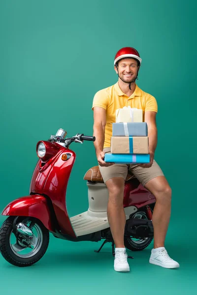 Cheerful delivery man in helmet holding wrapped presents near scooter on blue — Stock Photo