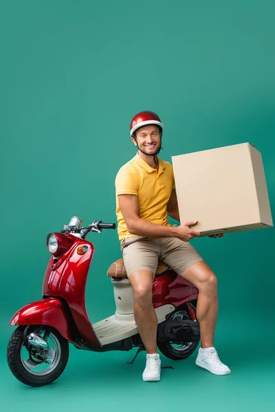 Fröhlicher Auslieferer im Helm hält großen Karton neben Roller auf blau — Stockfoto